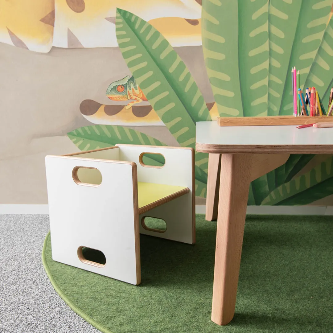 Kinderstuhl als Würfel mit Grifflöchern am Basteltisch – Papoq Sortiment Weißer Holzwürfelstuhl mit gelber Sitzfläche und ovalen Grifflöchern an einem Birkenholz-Basteltisch mit Buntstiften, tropische Wandbemalung im Hintergrund