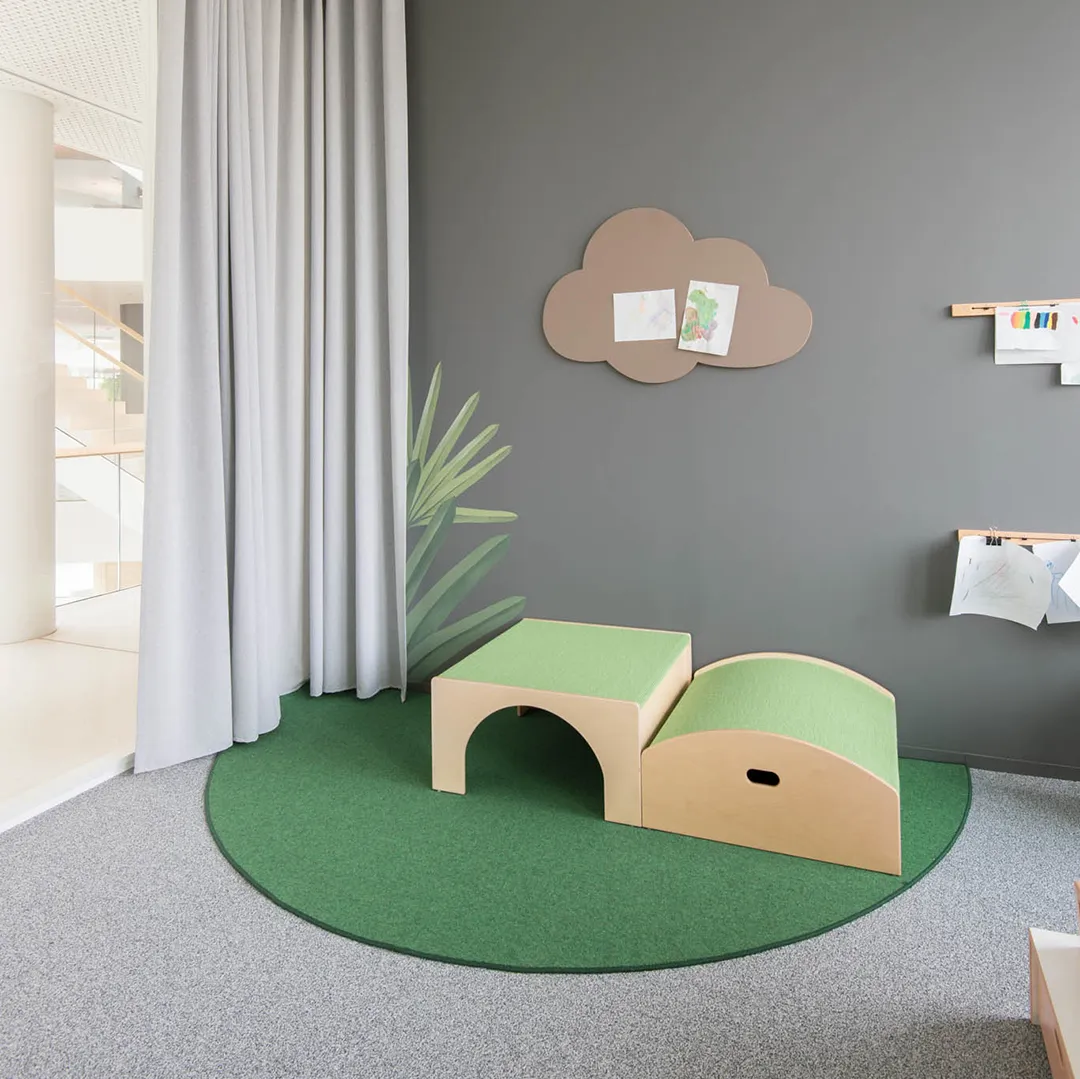 Kita-Rollenspielecke mit Tunnelmöbel und Wolkenpinnwand – Papoq Funktionsraum Grüne Spielmöbel mit Tunnelöffnung auf rundem grünem Teppich, darüber wolkenförmige Pinnwand mit Kinderzeichnungen an dunkelgrauer Wand