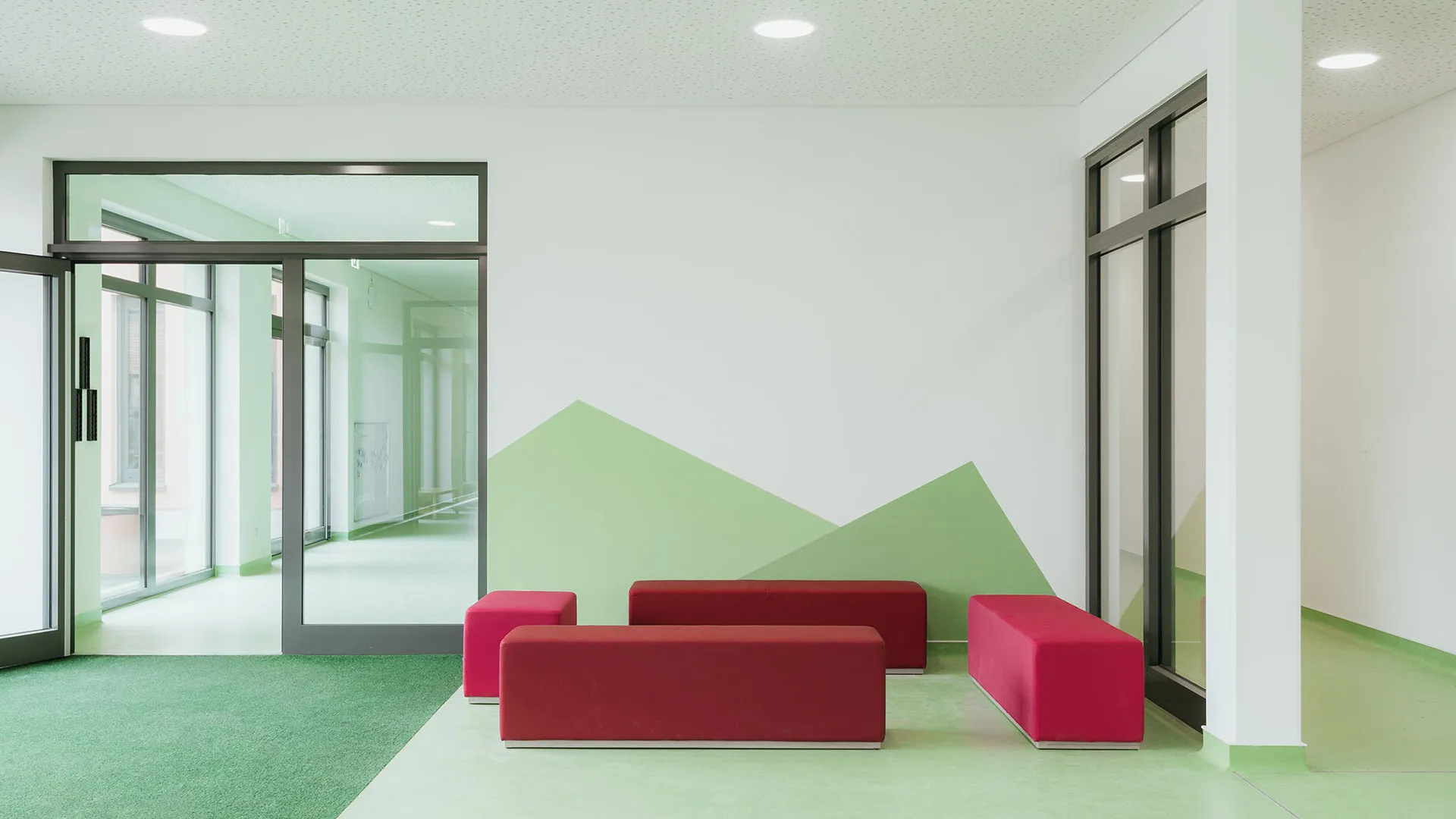 Kita-Eingangsbereich mit roten Sitzmöbeln und Wandgestaltung – Papoq Raumkonzept Heller Kita-Eingangsbereich mit drei roten Polsterbänken vor einer geometrischen Bergmotiv-Wandgestaltung in Hellgrün, grüner Bodenbelag