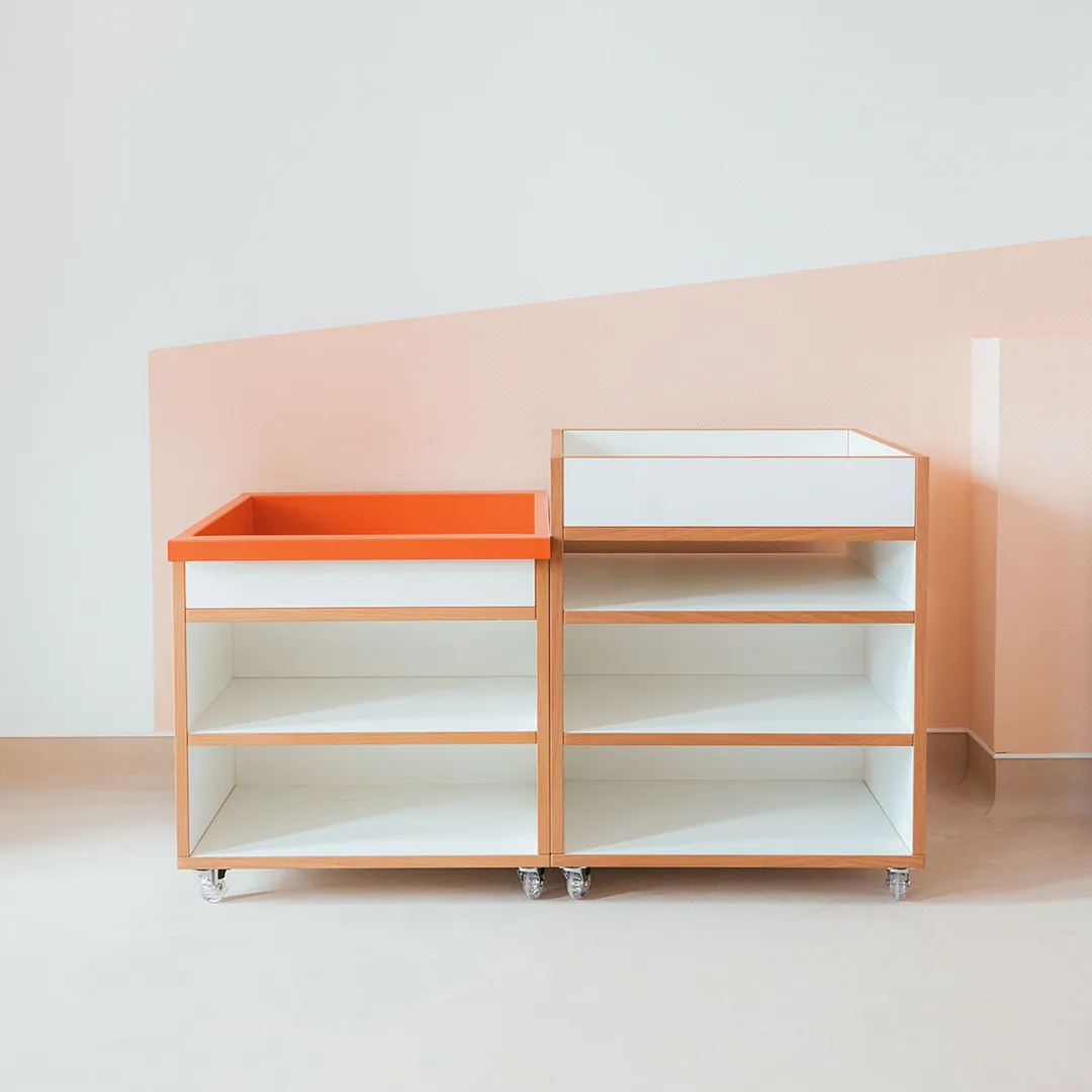 Fahrbare Wickelkommoden in Orange und Weiß – Papoq Funktionsraum Zwei fahrbare Wickelkommoden aus Birkenholz mit offenen Fächern, eine mit orangefarbener Wickelfläche, vor zweifarbiger Wandgestaltung in Apricot