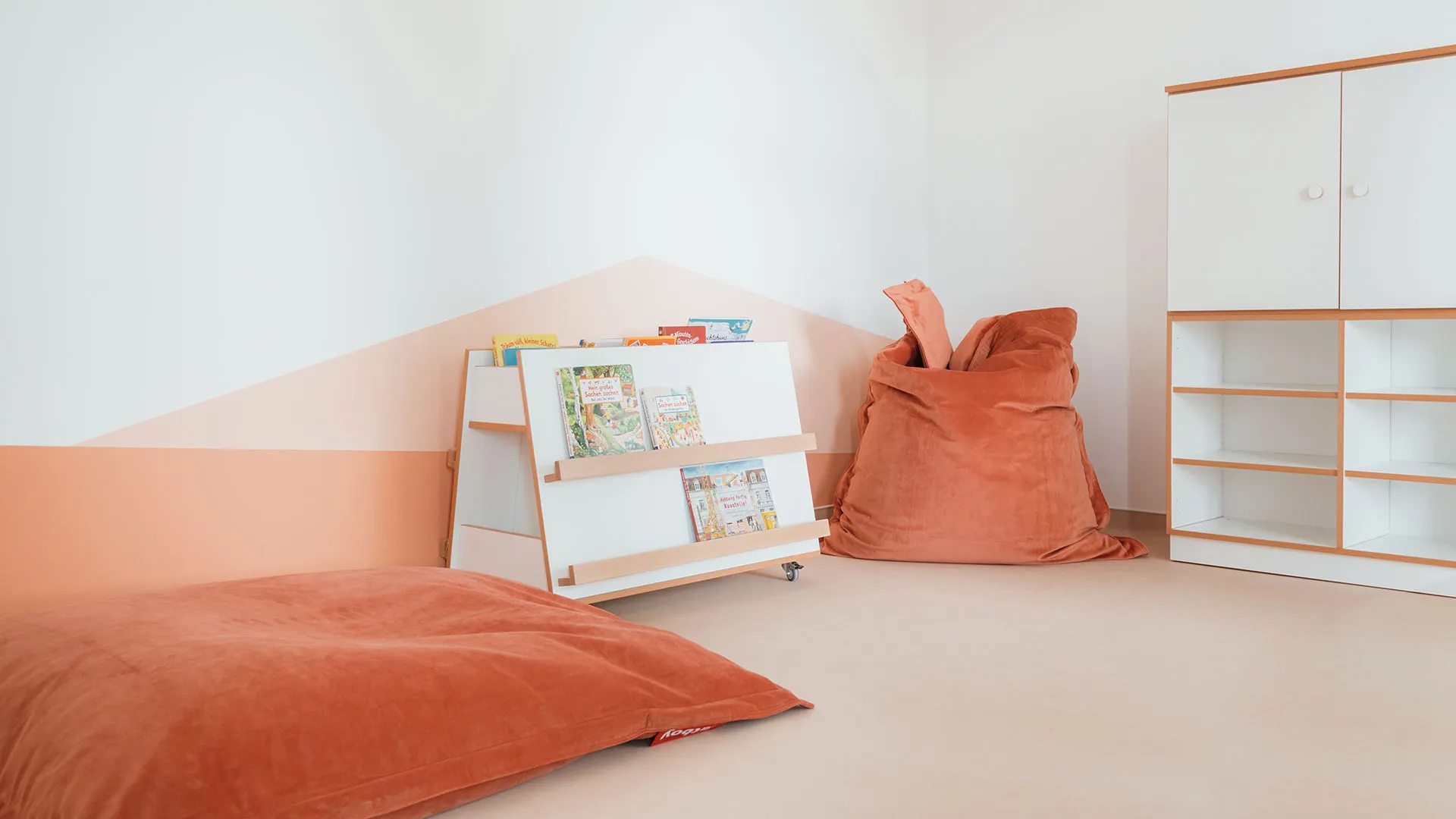 Kita-Leseecke mit Bücherregal und Sitzsäcken – Papoq Funktionsraum Helle Leseecke mit fahrbarem weißen Bücherregal, zwei terrakottafarbenen Sitzsäcken und Schrank, vor geometrischer Wandgestaltung in Apricot