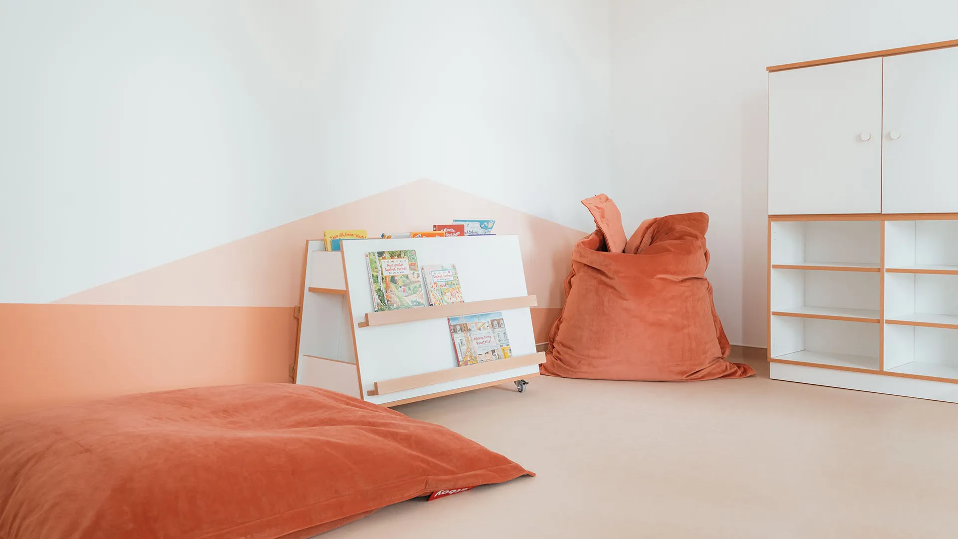 Kita-Leseecke mit Bücherregal und Sitzsäcken – Papoq Raumkonzept Helle Kita-Leseecke mit fahrbarem weißen Bücherregal, zwei terrakottafarbenen Sitzsäcken und geometrischer Wandgestaltung in Lachston