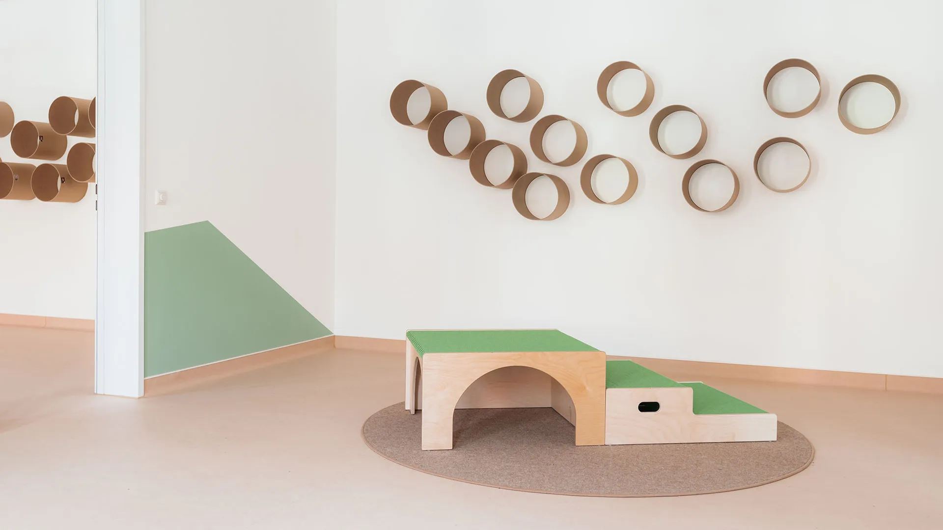 Kita-Schlafraum mit Spielpodest und Zylinderboxen – Papoq Raumkonzept Heller Kita-Raum mit grünem Spielpodest mit Rundbogen auf Filzteppich, zylindrischen Wandboxen in Sandton an weißer Wand, geometrische Wandgestaltung links