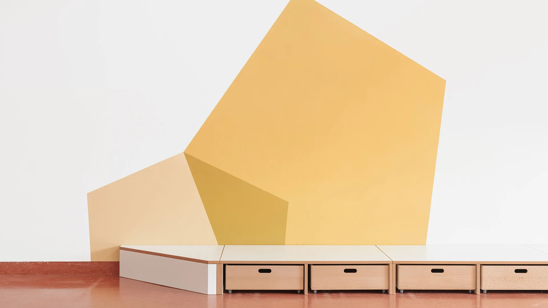 Kita-Podest mit geometrischer Wandgestaltung in Gelb – Papoq Raumkonzept Niedriges weißes Kita-Podest mit integrierten Holzschubladen vor großformatiger geometrischer Wandgestaltung in Gelb- und Cremetönen, terrakottafarbener Bodenbelag