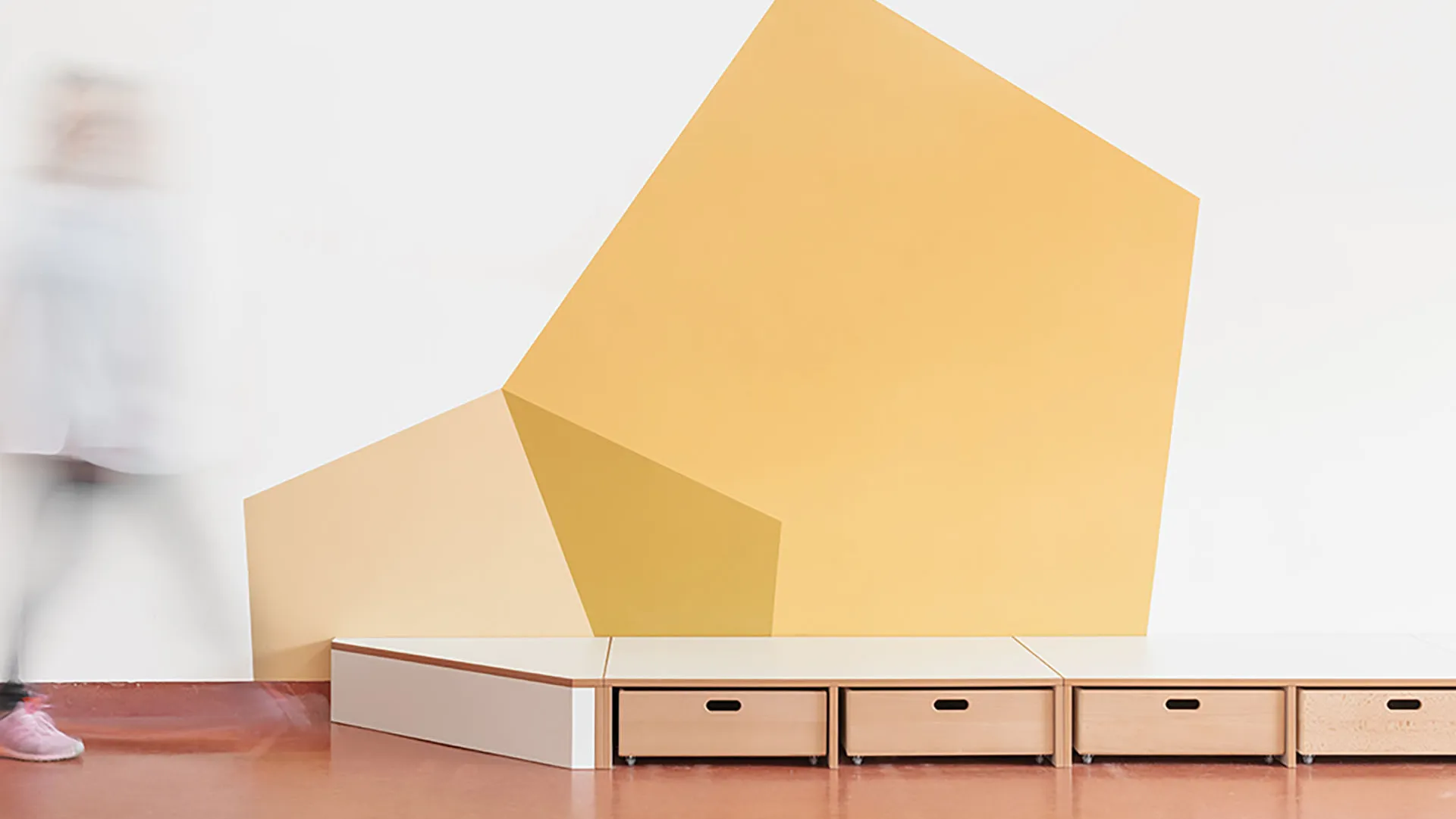 Kita-Podest mit geometrischer Wandgestaltung in Gelb – Papoq Funktionsraum Niedriges Podest mit Holzschubladen vor großer geometrischer Wandgestaltung in Gelb- und Cremetönen, Person in Bewegungsunschärfe im Hintergrund