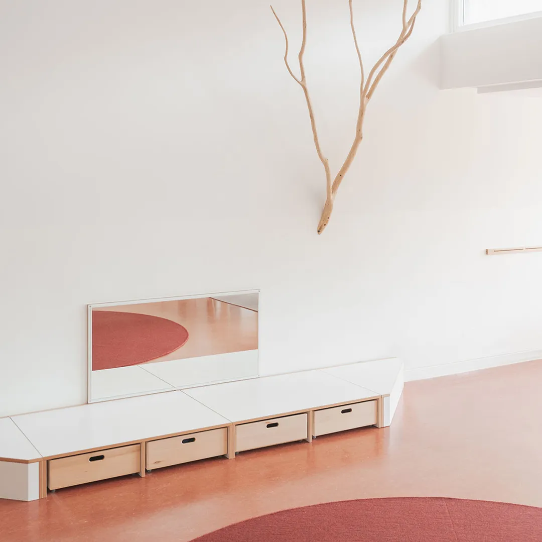 Kita-Podest mit Wandspiegel und Naturast – Papoq Raumkonzept Weißes Kita-Podest mit Schubladen und wandmontiertem Spiegel, darüber hängender Naturast, roter Rundteppich im Vordergrund, bordeauxroter Bodenbelag