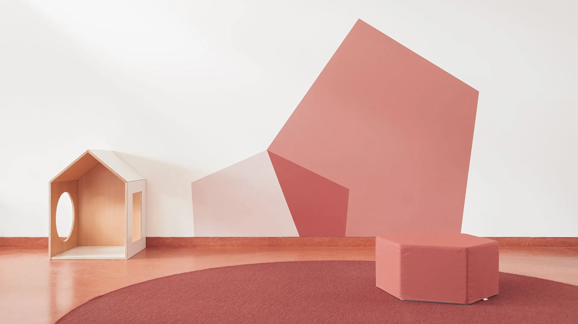 Kita-Spielhaus aus Holz vor Wandgestaltung in Rosé – Papoq Funktionsraum Kleines Holzspielhaus mit ovaler Aussparung neben einem rosafarbenen Sechseckhocker, vor geometrischer Wandgestaltung in Altrosa- und Mauvetönen