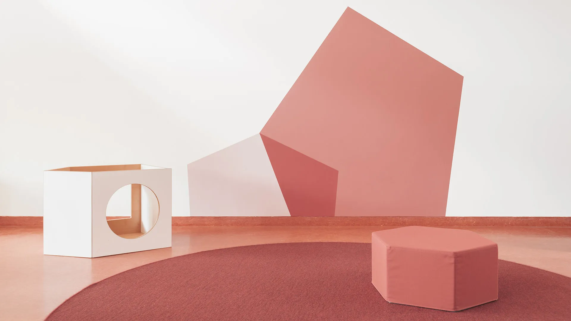 Kita-Spielmöbel vor geometrischer Wandgestaltung in Rosétönen – Papoq Raumkonzept Weißer Holzwürfel mit runder Aussparung und rosafarbener Sechseck-Polsterhocker auf bordeauxrotem Rundteppich vor großformatiger geometrischer Wandgestaltung in Altrosa und Mauve