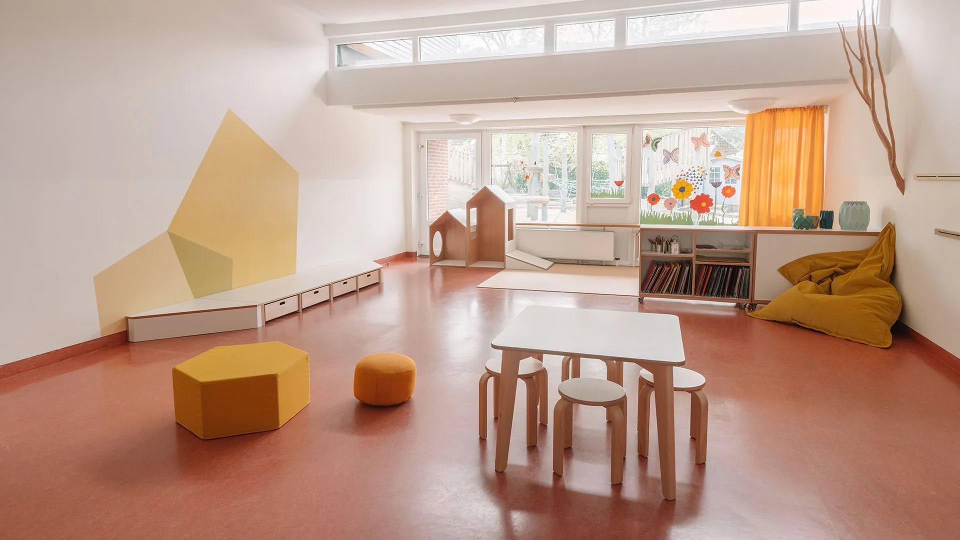 Kita-Gruppenraum mit Tisch, Hockern und Spielhaus – Papoq Raumkonzept Heller Kita-Gruppenraum mit weißem Kindertisch und Birkenholzhockern, Spielhaus aus Holz im Hintergrund, gelber Wandgestaltung, orangem Vorhang und Naturast an der Wand
