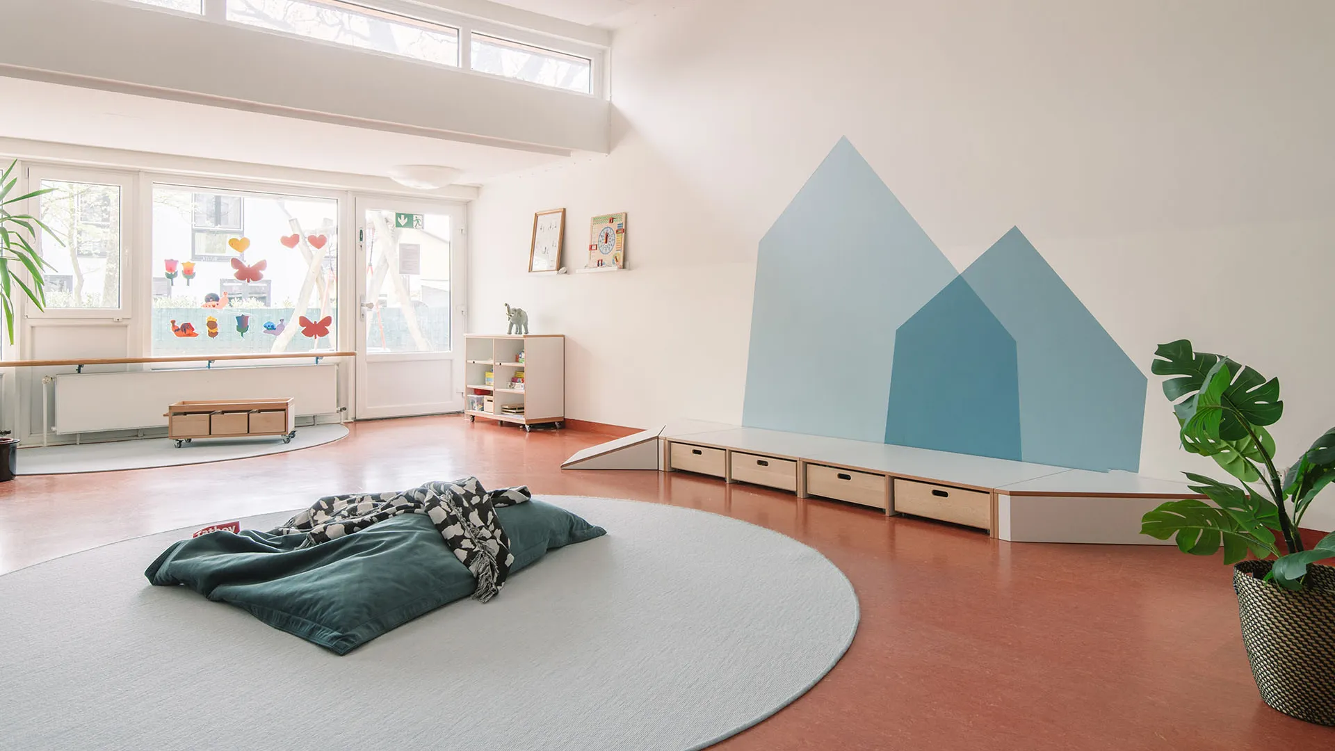 Kita-Gruppenraum mit blauer Haussilhouette und Sitzkissen – Papoq Raumkonzept Heller Kita-Gruppenraum mit geometrischer Wandgestaltung in Blautönen als Haussilhouette, Podest mit Schubladen und Rampe, grünem Sitzsack auf rundem Teppich im Vordergrund