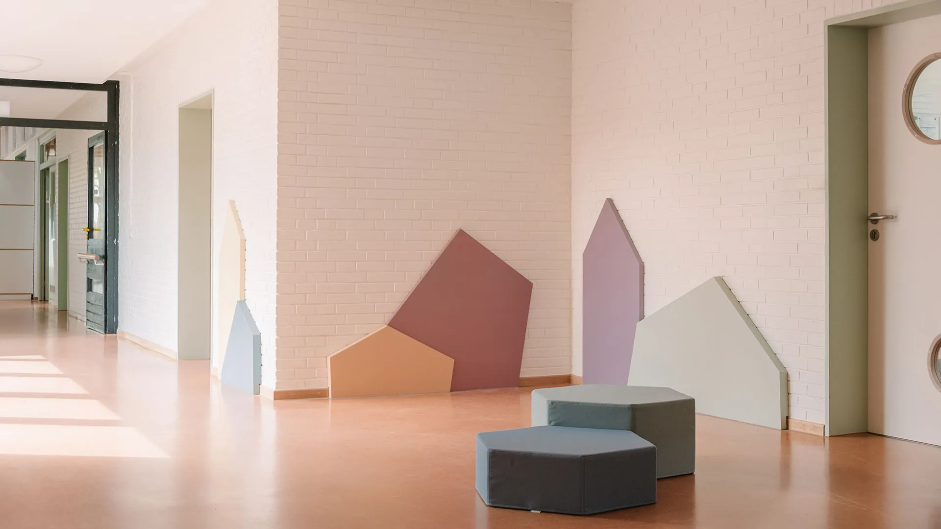 Heller Flur mit weißer Backsteinwand und fünf geometrischen Wandobjekten in Pastelltönen, davor zwei graue Sechseckhocker, lachsfarbener Bodenbelag