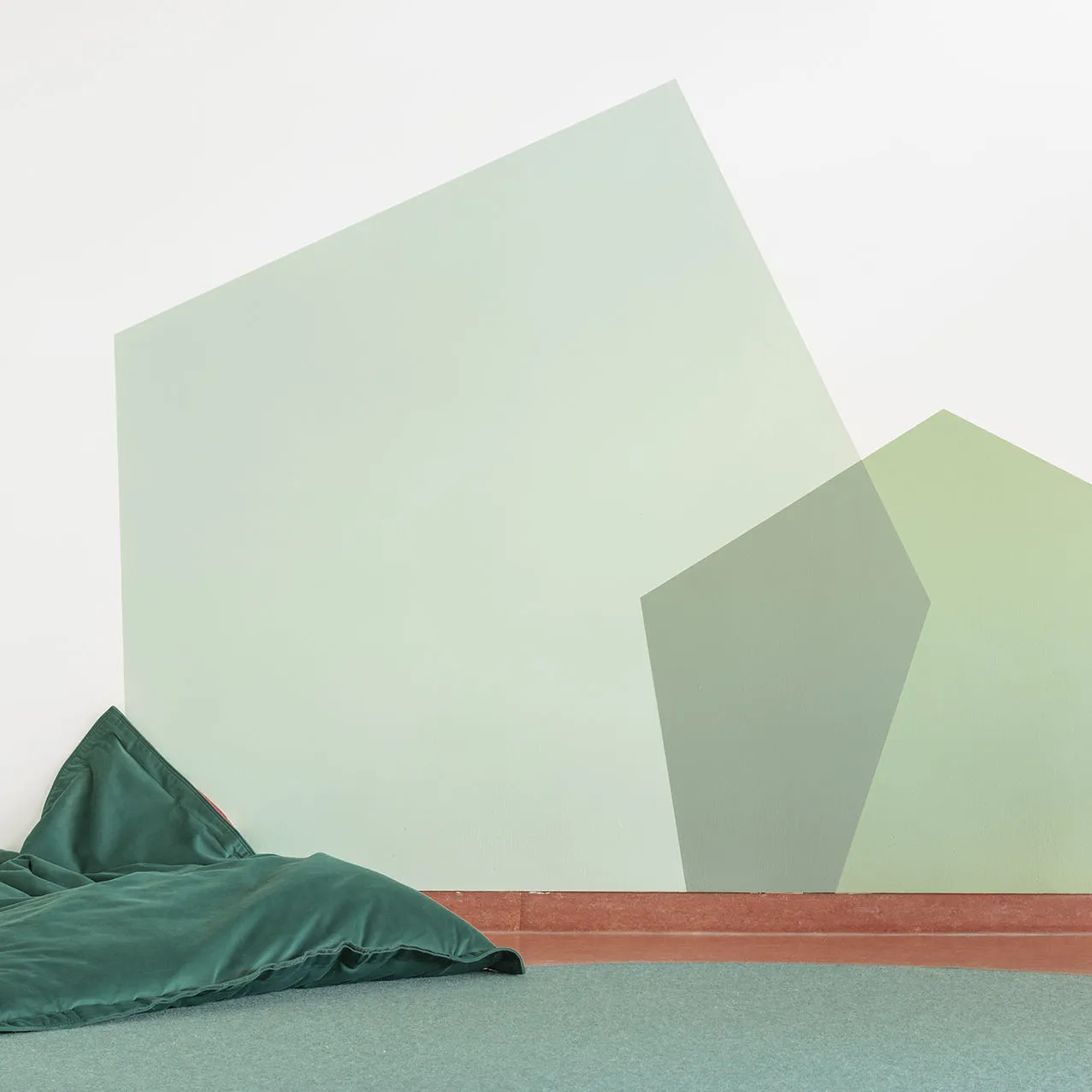 Ruheraum mit geometrischer Wandgestaltung in Grüntönen – Papoq Raumkonzept Detailaufnahme einer geometrischen Wandgestaltung aus überlappenden Flächen in Mintgrün und Salbei, davor dunkelgrüner Sitzsack auf petrolgrünem Teppich