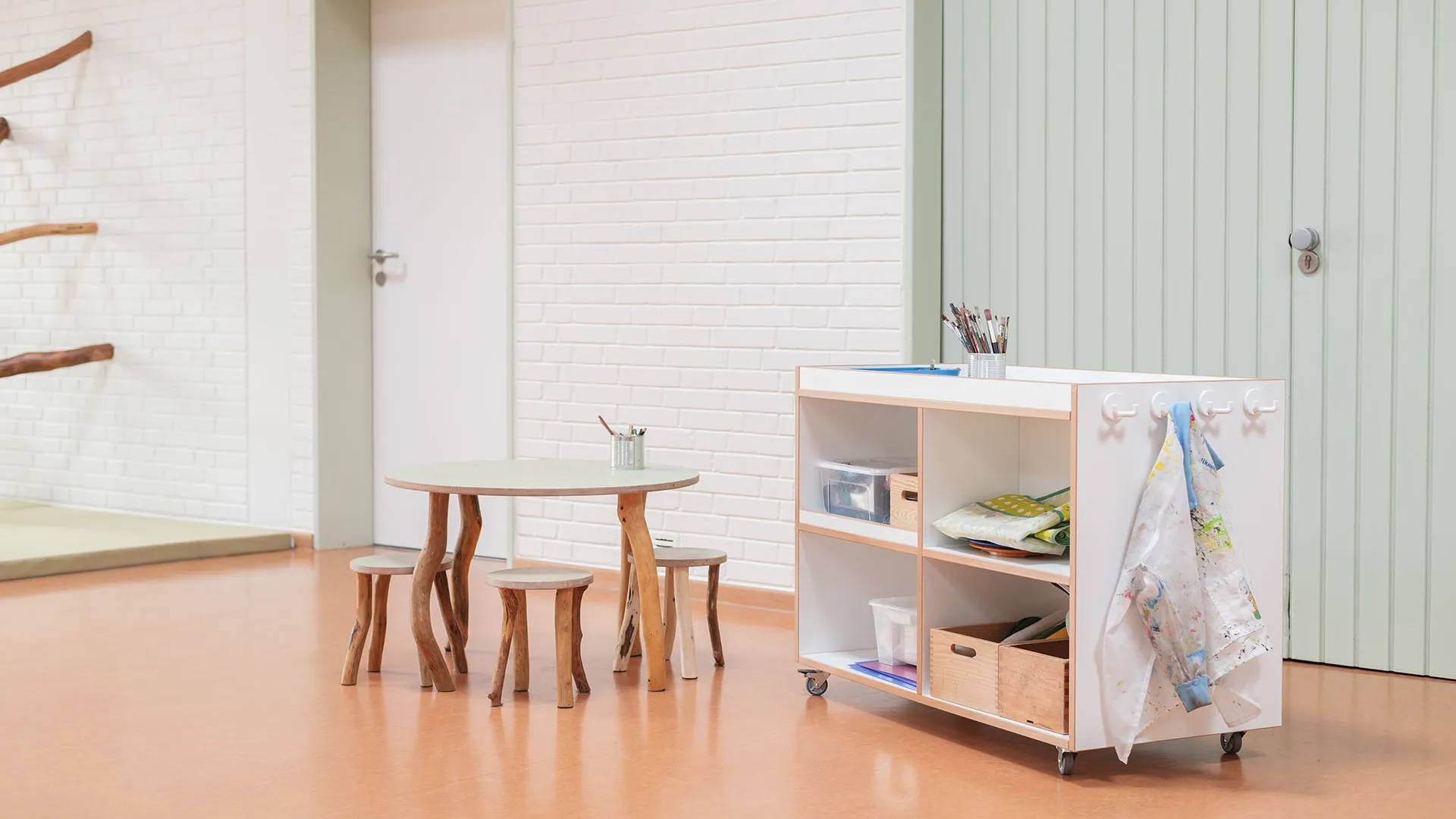 Kita-Atelier mit Maltisch und Materialwagen – Papoq Funktionsraum Runder Tisch mit Naturholzbeinen und passenden Hockern, daneben fahrbarer Materialwagen mit Schürzen und Aufbewahrungsboxen vor weißer Backsteinwand