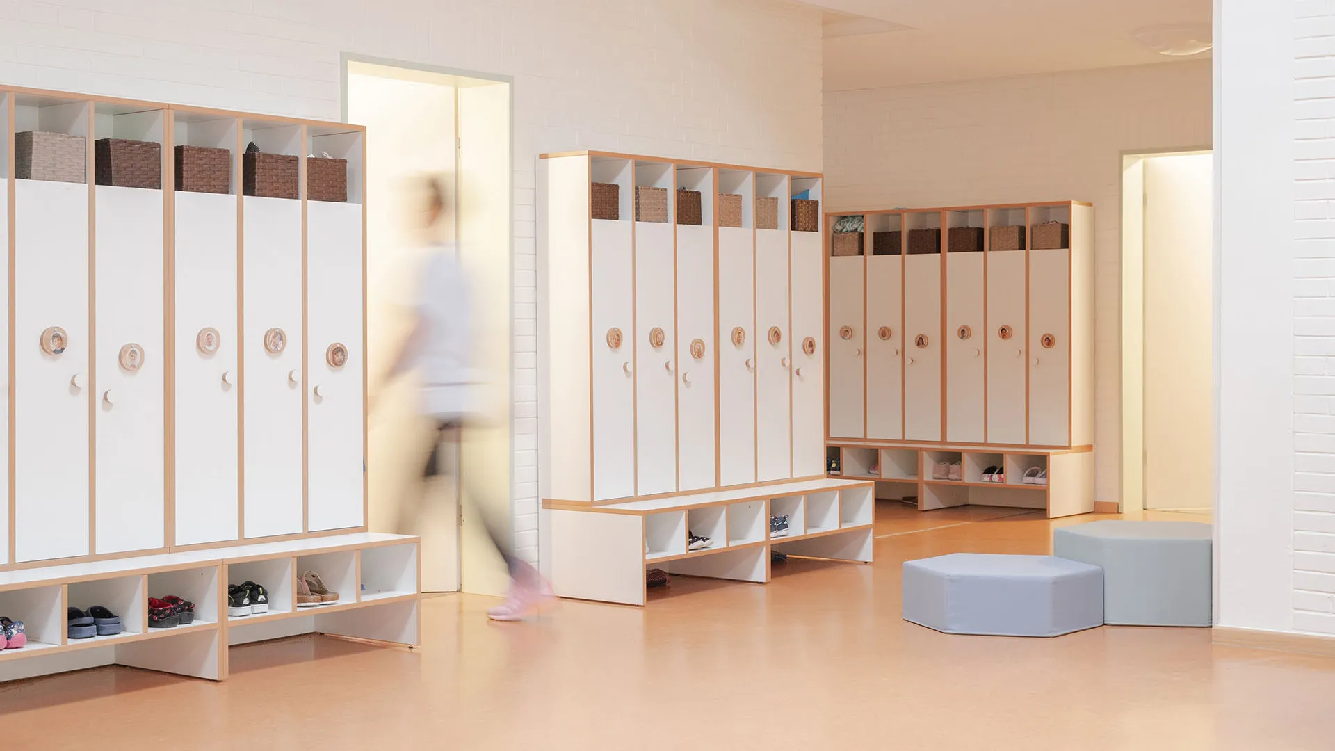 Drei weiße Garderobenschrankreihen mit Fotos der Kinder an den Türen, Körben oben und offenen Schuhfächern unten, Person in Bewegungsunschärfe