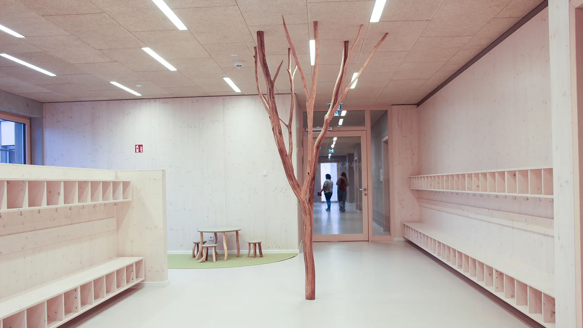 Heller Holzflur mit beidseitigen Garderobenfächern und Kleiderhaken aus hellem Holz, mittig ein Naturast als Raumteiler, kleiner Sitzbereich mit rundem Tisch im Hintergrund