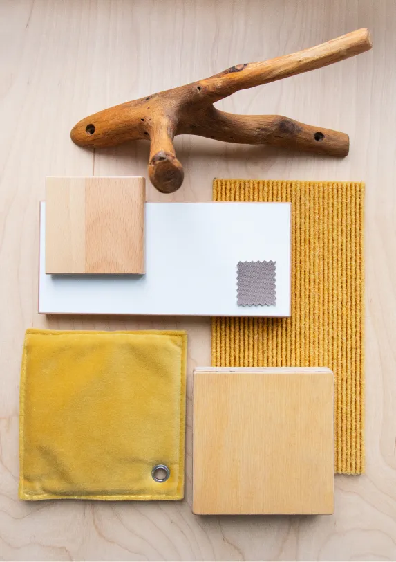 Materialboard in Gelbtönen für Kita-Raumkonzept – Papoq Planungsprozess Flatlay mit Materialmuster in Gelb- und Ockertönen: Holzwürfel, weißes Laminat, Samtstoff, Cordbezug, Teppichprobe und Naturholzast auf Birkensperrholz