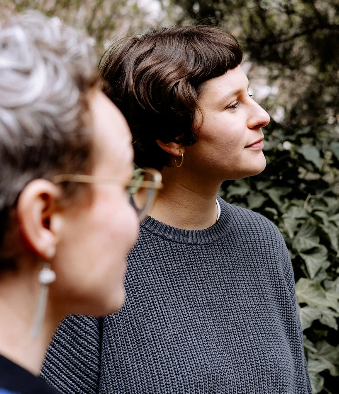 Lilia und Christine von Papoq im Gespräch Christine im Vordergrund unscharf, Lilia im Hintergrund scharf, beide im Profil vor grünem Laub