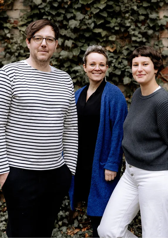 Das Papoq-Team: Lilia, Steffen und Christine Steffen, Lilia und Christine vom Papoq-Team stehen nebeneinander vor einer begrünten Wand