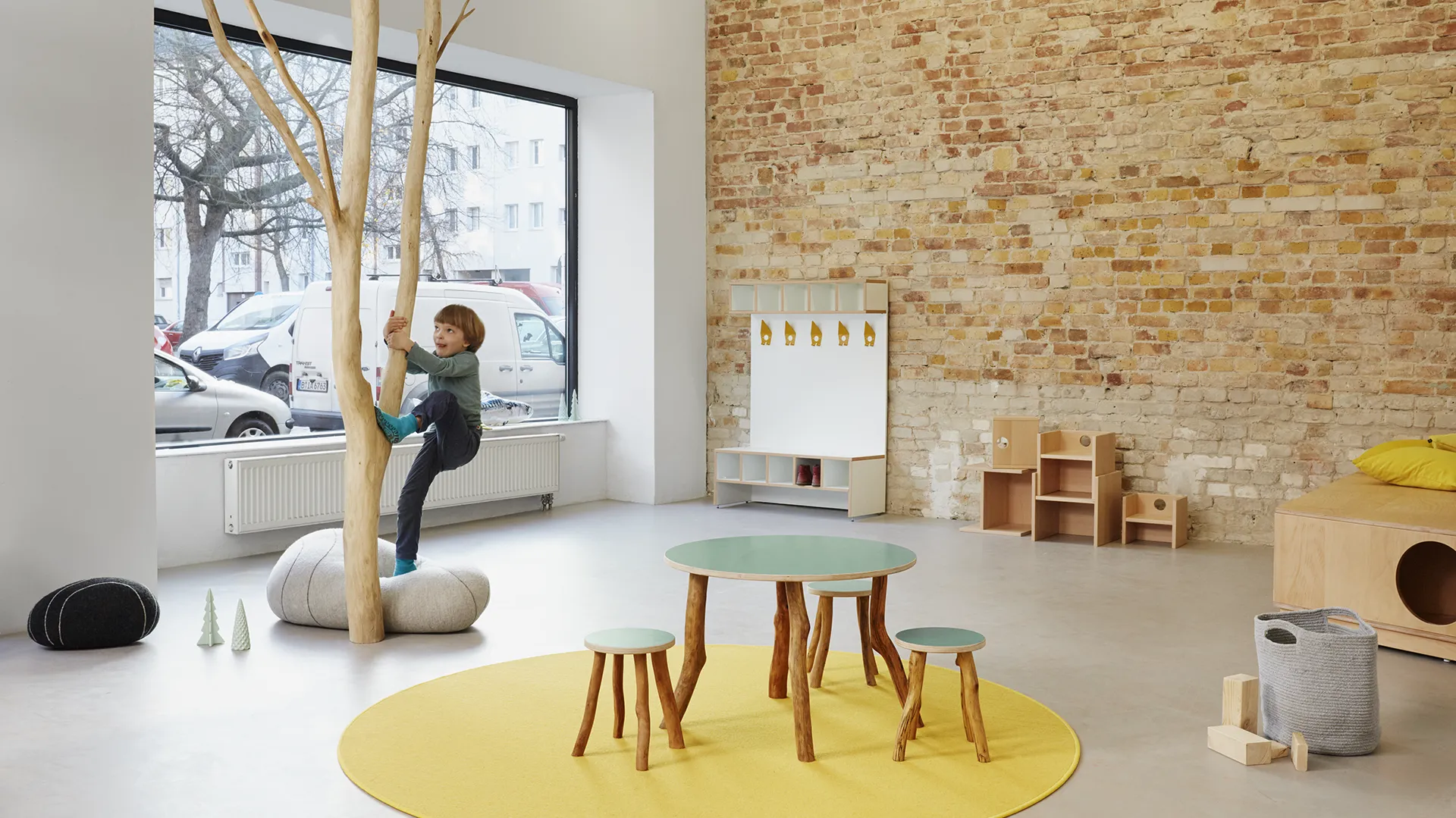 Multifunktionaler Kita-Raum mit Kletterast und Spielmöbeln – Papoq Funktionsraum Kind klettert an Naturholzast vor großem Fenster in multifunktionalem Kita-Raum, im Hintergrund Garderobenelement, Holzhocker und runder Tisch auf gelbem Teppich, Backsteinwand