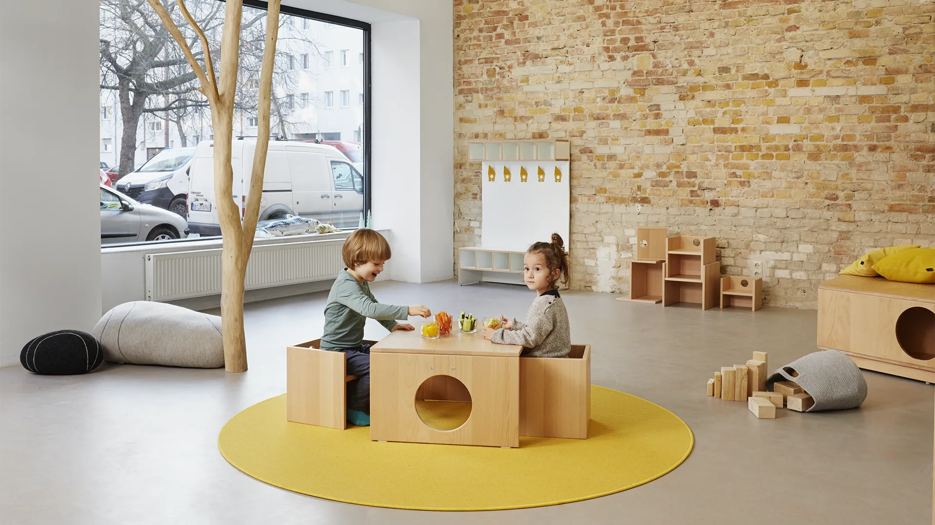 Zwei Kinder spielen gemeinsam am Kita-Tisch – Papoq Raumkonzept Zwei Kinder sitzen sich gegenüber an einem niedrigen Birkenholztisch mit runder Öffnung auf gelbem Rundteppich, Naturholzstamm und Backsteinwand im Hintergrund