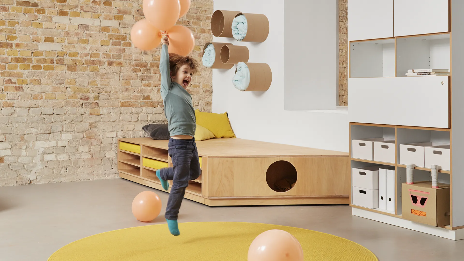 Kind springt jubelnd mit Luftballons vor einem Holzpodest mit runden Versteckröhren in einer Kita