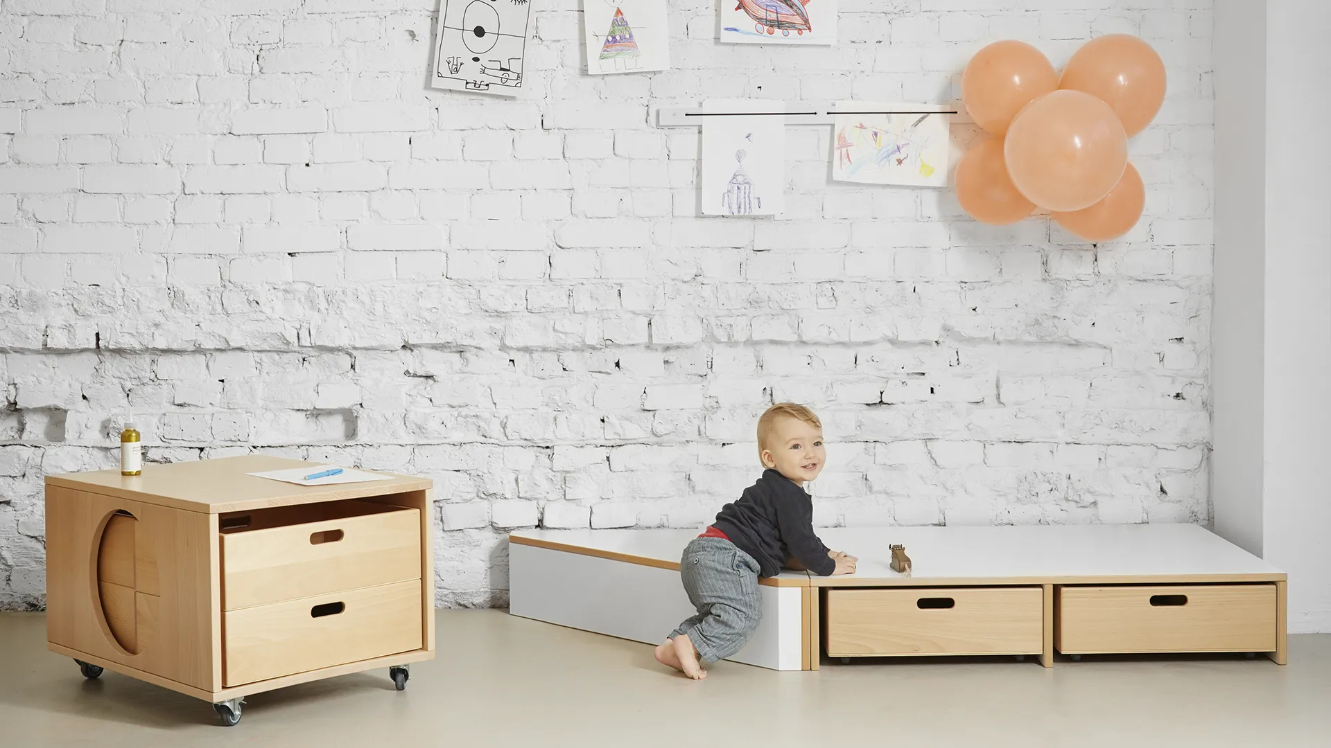 Kleinkind klettert auf Kita-Podest – Papoq Möbelkonzept Lachendes Kleinkind klettert barfuß auf ein weißes Kita-Podest mit Birkenholzschubladen, daneben fahrbares Regalmodul, Kinderzeichnungen und Luftballons an weißer Backsteinwand