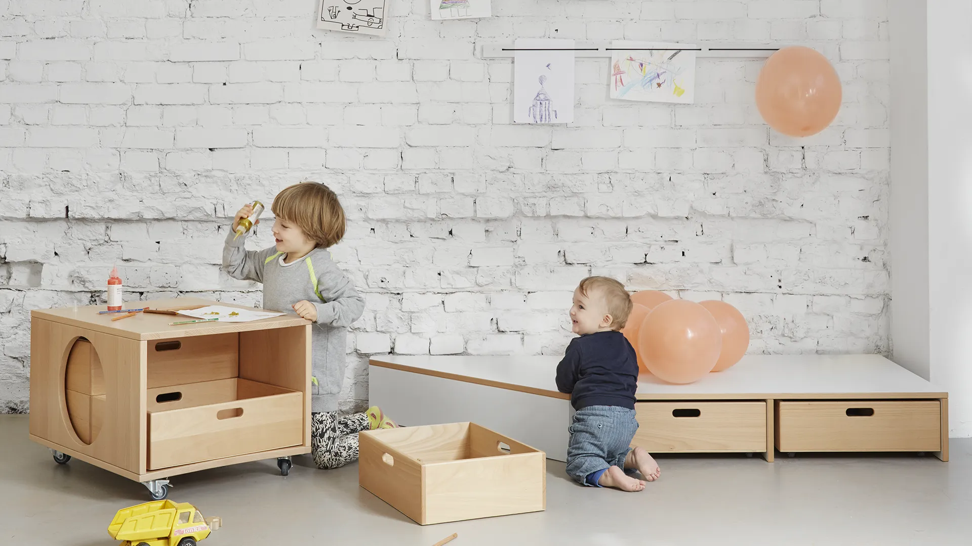 Zwei Kinder spielen mit Kita-Aufbewahrungsbox – Papoq Funktionsraum Älteres Kind spielt mit fahrbarer Holzbox mit runder Aussparung und Schubladen, Kleinkind krabbelt zu Luftballons an Podest mit Schubladen
