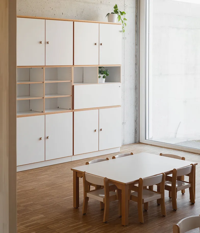 Heller Kita-Gruppenraum mit modularer Stauraumwand aus weißen Schränken und offenen Holzregalen, niedrigem Esstisch und kindgerechten Holzstühlen von papoq.