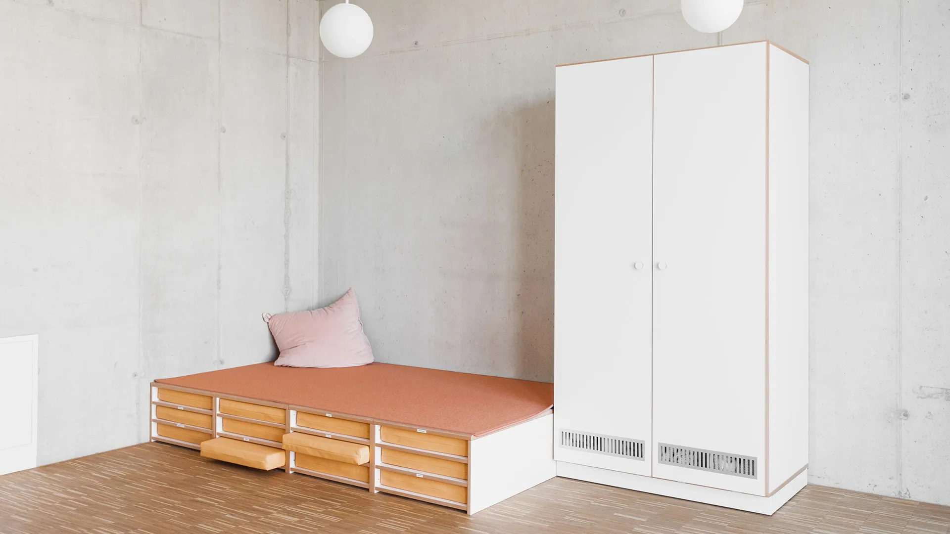 Kita-Schlafpodest mit Liegekissen und geschlossenem Schrank – Papoq Ruheraum Niedriges Schlafpodest mit terrakottafarbenem Bezug, rosa Kissen und integrierten Schubkästen, daneben geschlossener weißer Zweitürenschrank, Sichtbetonwände, Holzboden