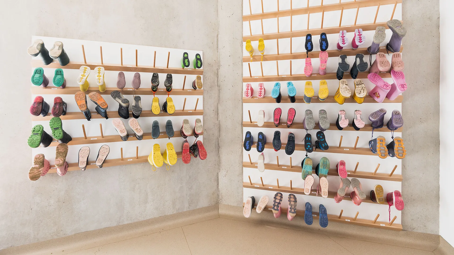 Zwei wandmontierte Holzschuhregale mit Holzstiften zum Aufhängen von Kinderschuhen, dicht bestückt mit bunten Schuhen und Gummistiefeln, Sichtbetonwände