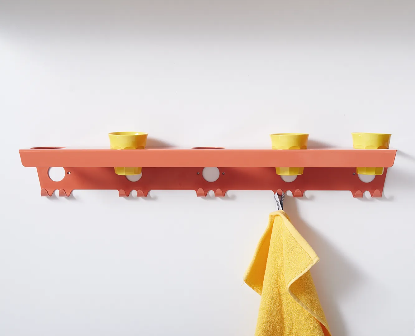 Orangefarbene Wandleiste für Kitas mit gelben Zahnputzbechern und Handtuchhaken – ein gelbes Handtuch hängt an einem Haken
