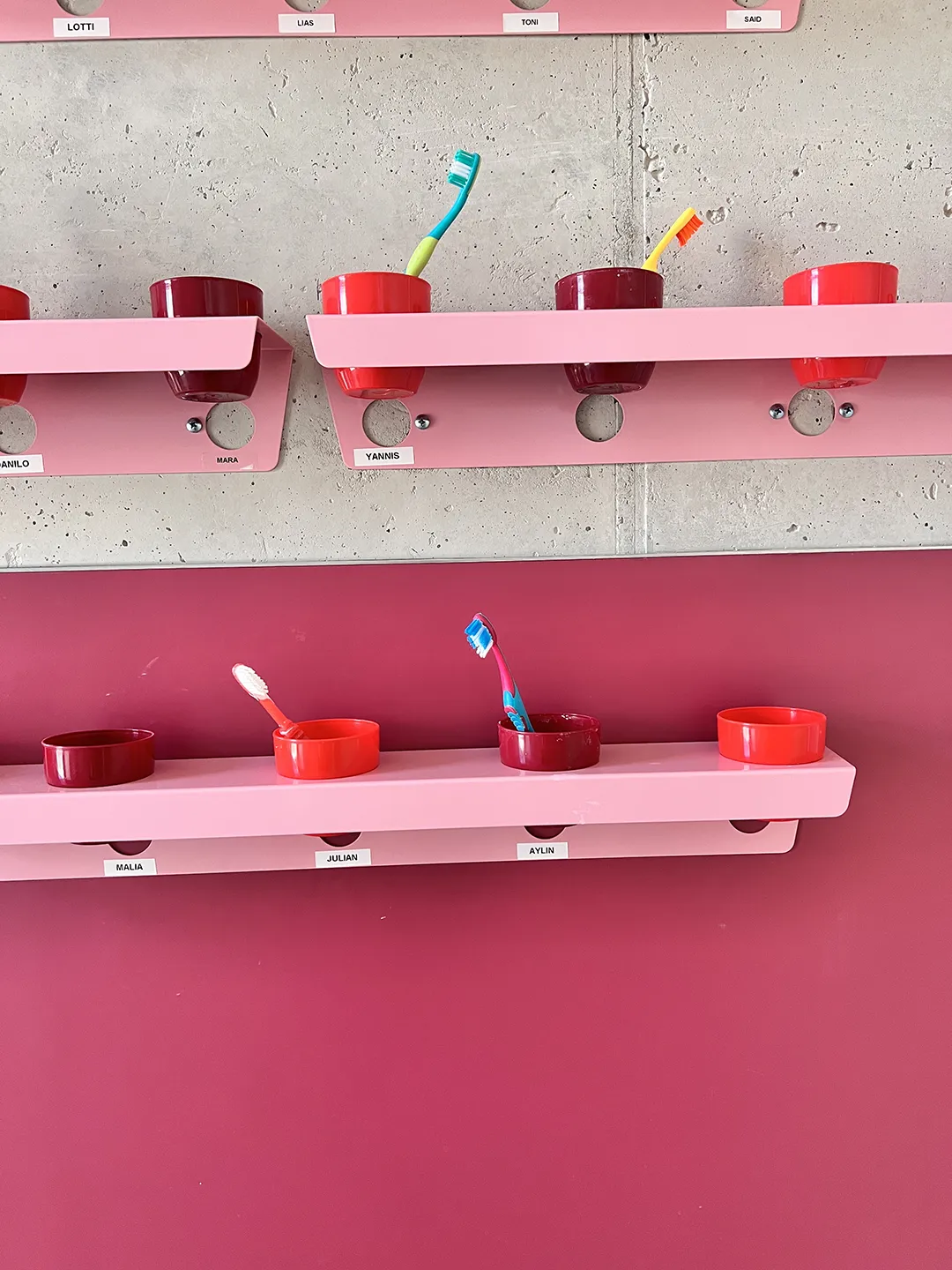 Pinke Wandregale mit roten Zahnputzbechern und namentlicher Beschriftung für Kinder in einer Kita