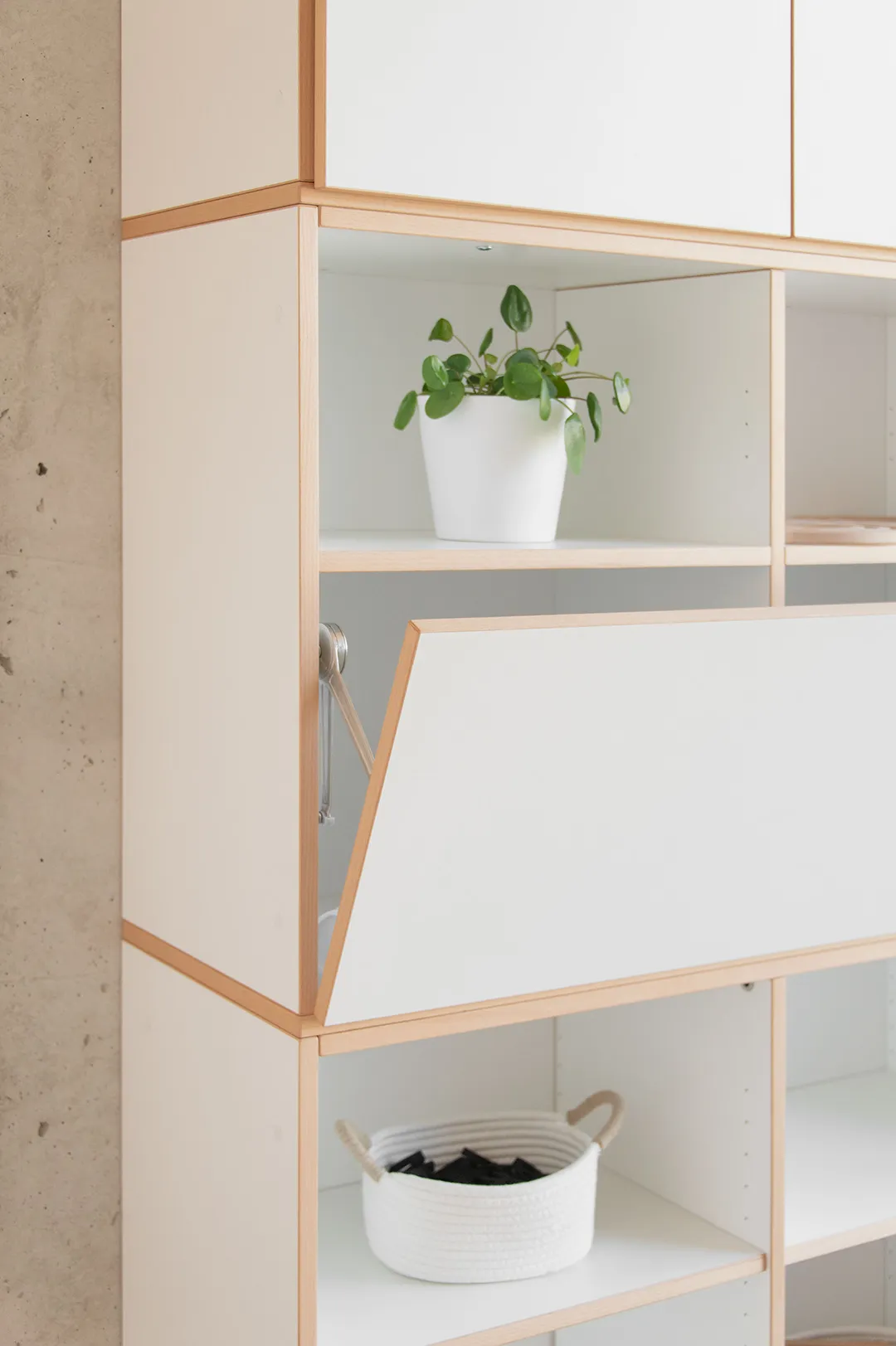 Modulares weißes Kita-Regal mit Buchenholzrahmen – Klapptür halb geöffnet, Pflanze und Korb in den Fächern
