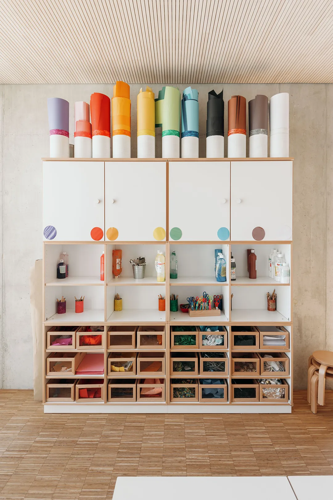 Weißes Kita-Regalsystem mit bunten Papierrollen obenauf, Holzboxen mit durchsichtigen Fronten und beschrifteten Fächern für den Atelierbereich